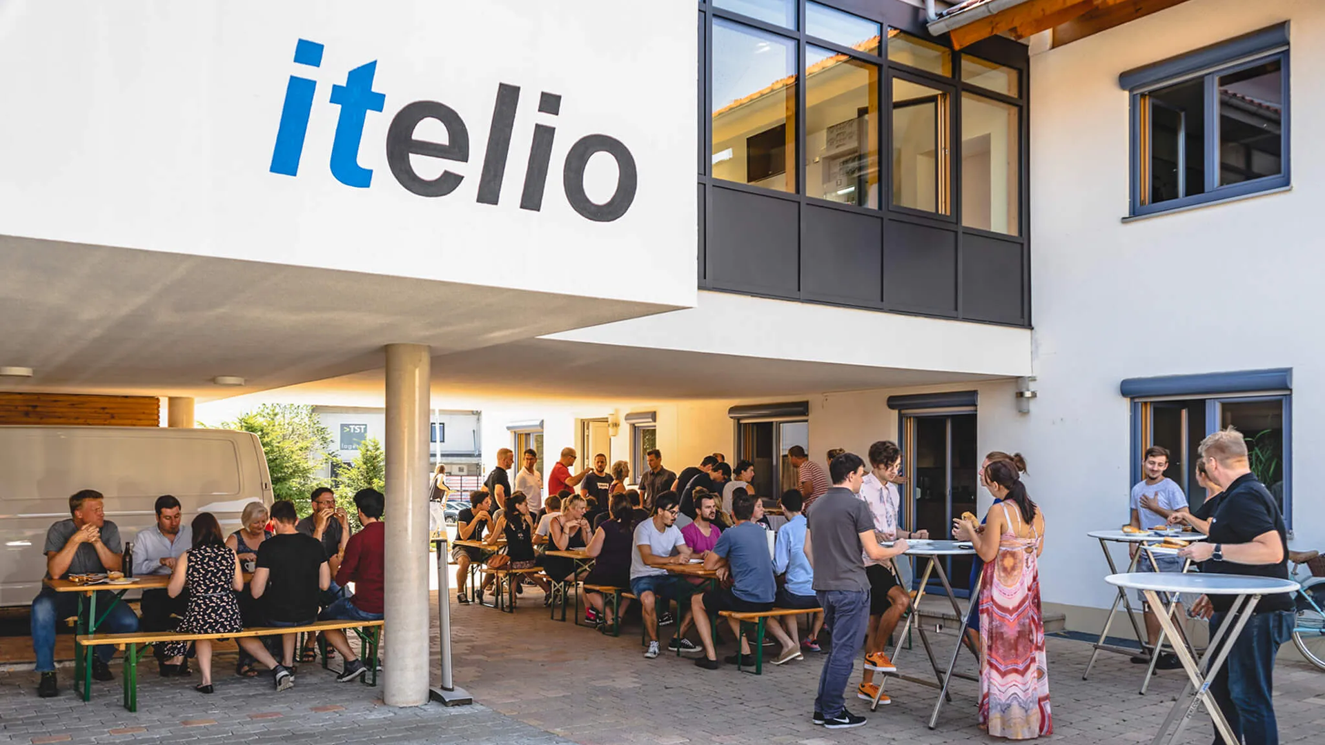 Zahlreiche Mitarbeitende sitzen an Biertischgarnituren und stehen an Stehtischen im Freien vor dem Firmengebäude mit großem itelio-Schriftzug