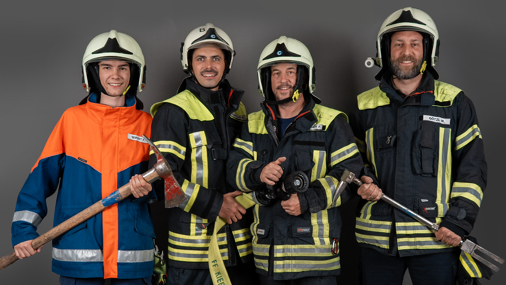 Vier Feuerwehrleute in Schutzkleidung und Helmen posieren mit Axt, Strahlrohr und Einreißhaken vor grauem Studiohintergrund