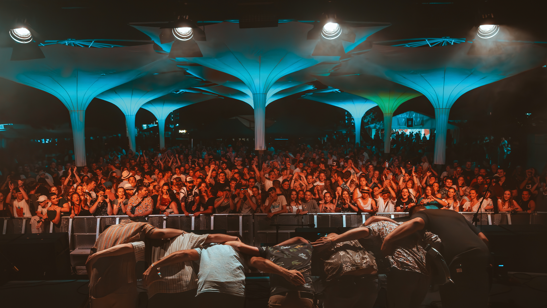 View from the stage at the Kurpark Open Air Festival in Kiefersfelden – musicians take a bow in front of a packed crowd under turquoise-lit canopy structures at night.