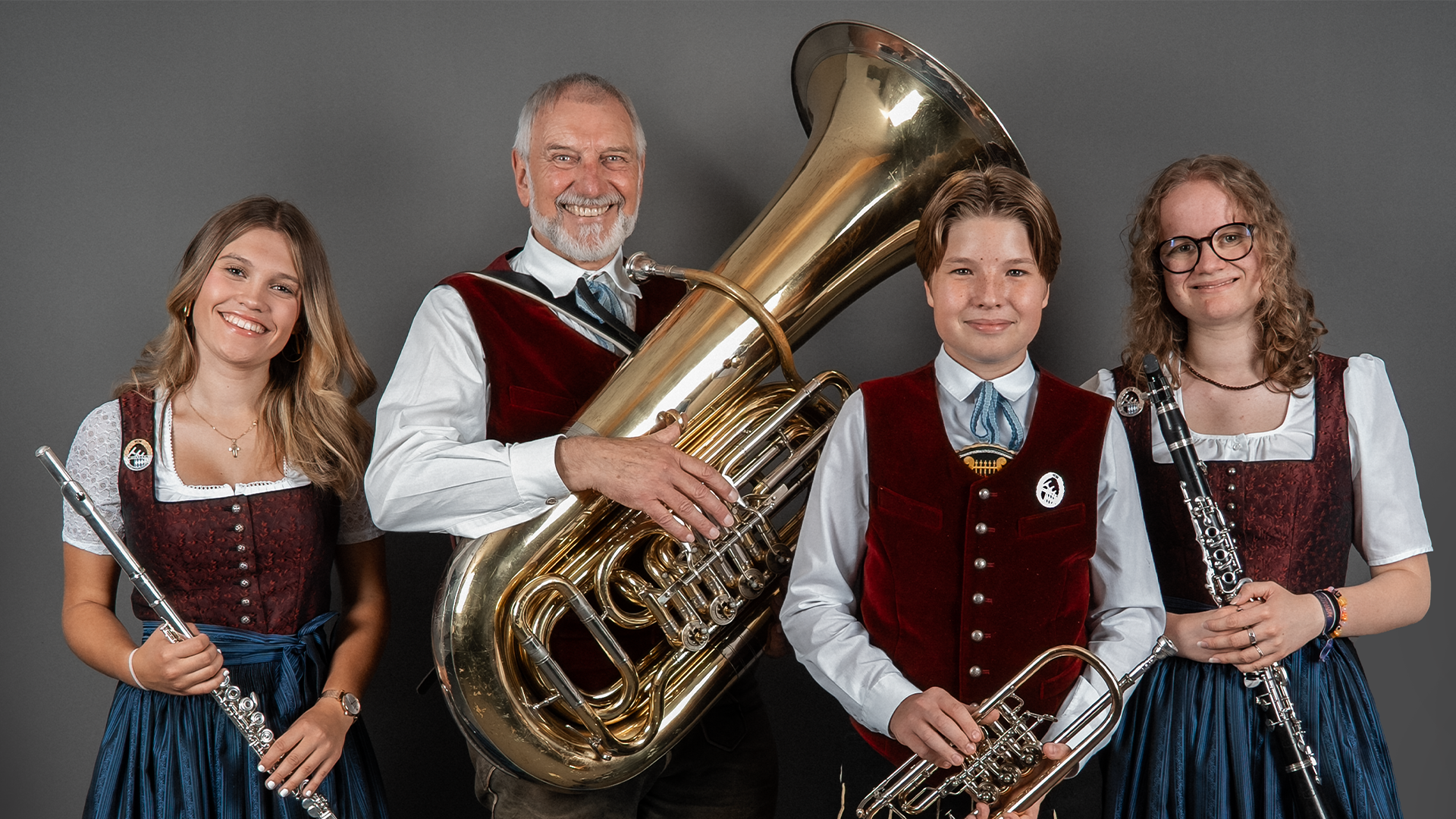 Four members of the Musikkapelle Kiefersfelden brass band in traditional Bavarian Tracht against a grey backdrop – from left: a young woman with a flute, an older gentleman with a tuba, a teenager with a trumpet and a young woman with a clarinet.