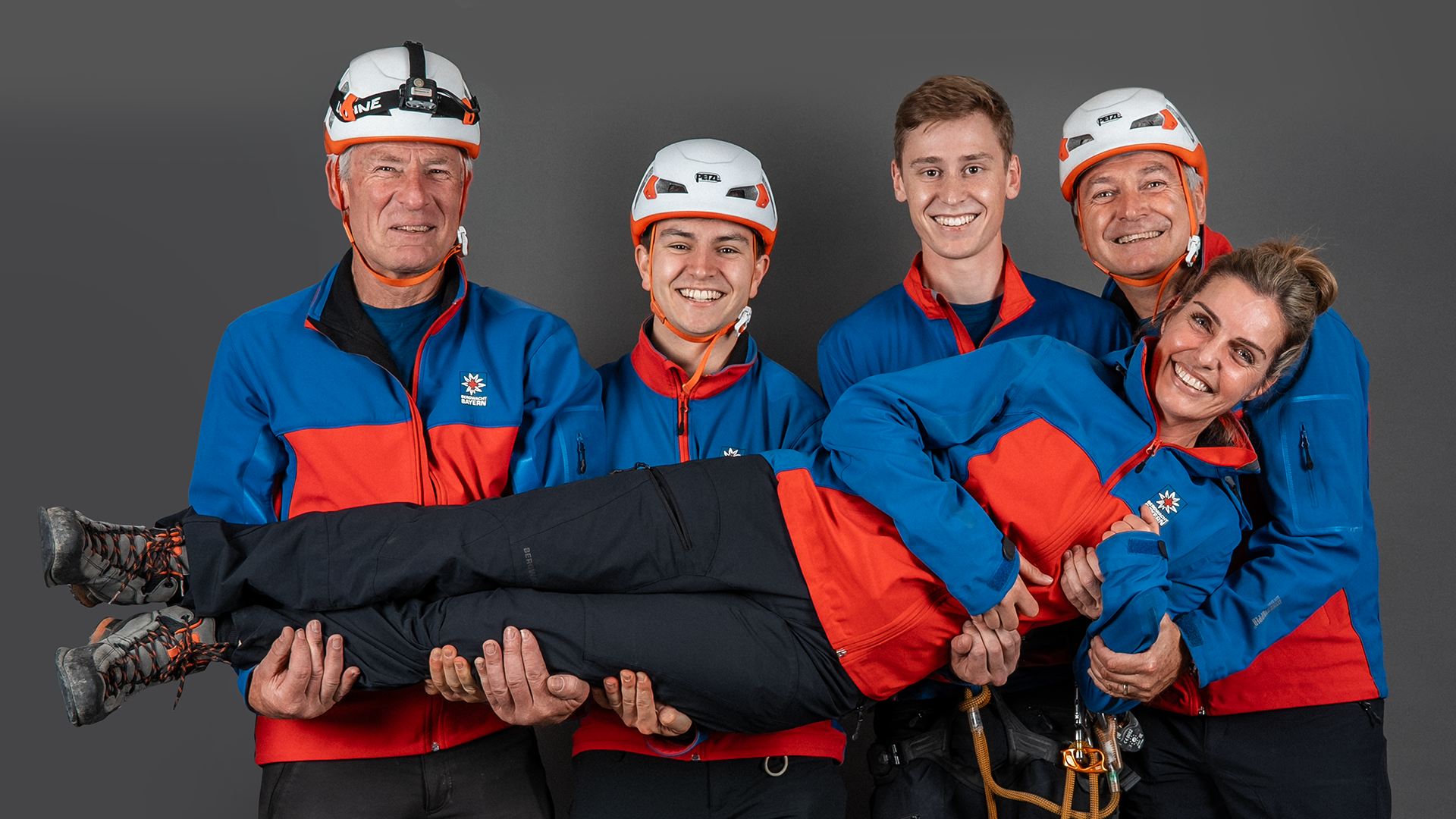 Five members of Bergwacht Bayern mountain rescue in blue-and-red gear with Petzl helmets against a grey backdrop – four carry a teammate in their arms, all laughing in a playful pose.
