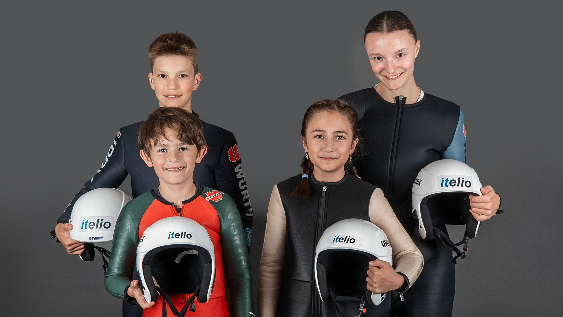 Four young ski jumpers from the Wintersportverein Kiefersfelden in jumpsuits against a grey backdrop, each holding a white helmet with the itelio logo.